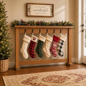 May include: Six Christmas stockings hanging from a wooden console table. The stockings feature various designs including cream cable knit, red and black buffalo plaid with a paw print, solid dark green knit, and a red and white snowflake pattern.