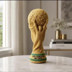 Könnte beinhalten: Eine goldgelbe Häkeltrophäe, die eine stilisierte menschliche Figur darstellt, die einen Globus hochhält. Der Sockel ist mit zwei horizontalen grünen Streifen akzentuiert und trägt den schwarzen Sticktext FIFA WORLD CUP 18K GOLD.