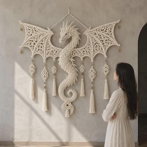 May include: A large off-white macrame wall hanging shaped like a dragon with intricate knotted wings, a textured serpentine body, and dangling tassels. The design features metallic gold beads integrated throughout the structure and along the edges.
