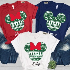 May include: Three short-sleeve t-shirts in red, navy blue, and white. Each shirt features a green graphic of a mouse-shaped ornament with a red bow, decorated with white reindeer, pine trees, and snowflakes. The shirts have text labels reading Mommy, Daddy, and Emily.
