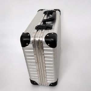 May include: A rectangular silver aluminium hard-shell briefcase featuring a ribbed exterior design, black plastic corner protectors, a black top handle, and black locking latches. The case is constructed with visible metal rivets along the frame.
