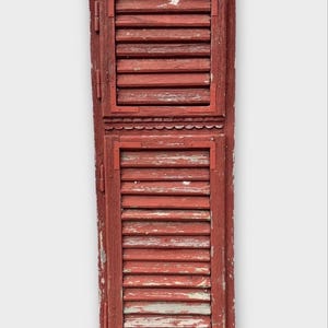 Könnte beinhalten: Ein hoher, rechteckiger Fensterladen aus Holz mit einer verwitterten, abgenutzten roten Lackierung. Er besteht aus zwei Abschnitten mit horizontalen Lamellen, die von Metallbeschlägen eingerahmt sind, und einer dekorativen Bogenkante.