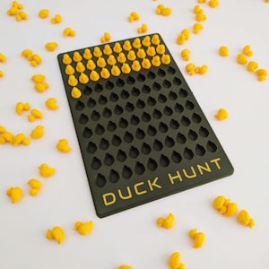 Op de afbeelding: Een rechthoekig zwart dienblad met een raster van kleine inkepingen en de tekst DUCK HUNT in het geel gedrukt. Het dienblad is gedeeltelijk gevuld met kleine gele eendvormige figuurtjes, met extra losse gele eendjes verspreid rondom.