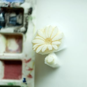 Könnte beinhalten: Ein weißer Schaumstoffstempel mit einem Gänseblümchen-Motiv, das ein kreisförmiges Zentrum und strahlenförmige Blütenblätter aufweist. Der Stempel hat einen rechteckigen Griff und liegt neben einer Palette mit verschiedenen Aquarellfarben.