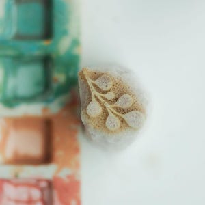 Op de afbeelding: Een kleine, cilindervormige rubberen stempel met een delicaat, geëtst botanisch bladontwerp op de voorzijde. De stempel ligt naast een verticale strook aquarelverfblokjes in tinten blauwgroen, perzik en gedempt rood met inktresten.