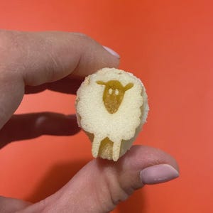 Puede incluir: Un pequeño bizcocho redondo de color beige que presenta un gráfico detallado de color marrón oscuro de una oveja en su superficie. El pastel tiene una textura suave y porosa y un borde festoneado distintivo que perfila la forma del cuerpo lanudo.