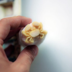 Pode incluir: Um pequeno recorte de espuma texturizada amarelo-claro em forma de caranguejo, com um corpo central oval e oito pernas articuladas. A forma de caranguejo está fixada numa base cilíndrica de espuma branca e macia, criando um elemento decorativo tridimensional.