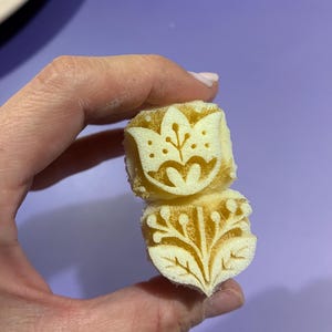 Può includere: Due timbri in schiuma gialla impilati con intricati disegni intagliati. Il timbro superiore mostra un fiore stilizzato simile a un tulipano con punti, mentre il timbro inferiore presenta un motivo botanico con foglie e bacche.