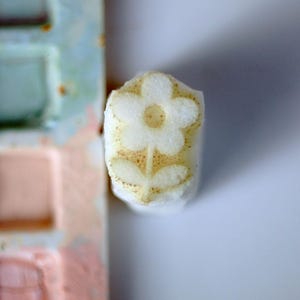 Puede incluir: Un sello rectangular blanco con un diseño de flor en relieve con cinco pétalos y dos hojas. La superficie del sello está ligeramente espolvoreada con un pigmento dorado amarronado, resaltando el patrón floral en relieve sobre el material blanco.
