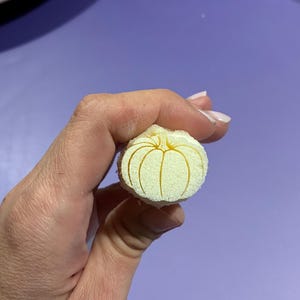 Könnte beinhalten: Ein kleiner, runder, cremeweißer Schwammstempel mit einem eingravierten Kürbismotiv auf der flachen Oberfläche. Der Stempel besteht aus einem texturierten, porösen Material und zeigt eine einfache, lineare Kürbisform mit Segmenten.