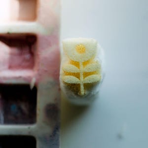 May include: A pale yellow stamp featuring a stylised flower design with a circular centre and two pairs of leaves on a vertical stem. The stamp is positioned next to a rectangular, multi-compartment tray with faded pink and brown paint residue.