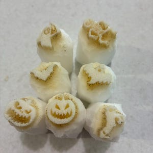 Könnte beinhalten: Ein Set aus sechs weißen, zylindrischen Zuckerwürfeln, die jeweils ein individuelles, bräunliches Halloween-Motiv auf der Oberseite aufweisen, darunter ein Kürbisgesicht, ein Geist und eine Fledermaus, angeordnet in einer Gruppe.