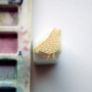 Op de afbeelding: Een kleine, beige, vogelvormige schuimrubberen stempel met een getextureerd, poreus oppervlak. De stempel staat verticaal naast een rechthoekig plastic palet met verschillende kleine, vierkante vakjes gevuld met diverse kleuren gedroogde aquarelverf.