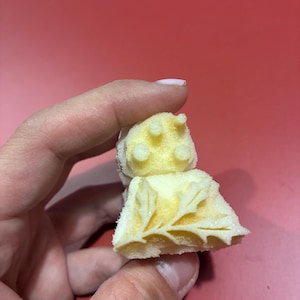 May include: A small, pale yellow, porous sponge-like object with a textured surface. The object features a carved leaf pattern on one side and three small, raised circular protrusions on the top section, showing a soft, foam-like material.