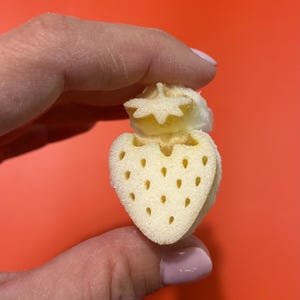 Könnte beinhalten: Ein kleiner, heller, poröser Schwamm in Erdbeerform. Der Schwamm hat eine strukturierte Oberfläche mit kleinen, gleichmäßig verteilten Vertiefungen, die Samen darstellen, sowie einen markanten blattförmigen oberen Teil aus weichem Schaumstoff.