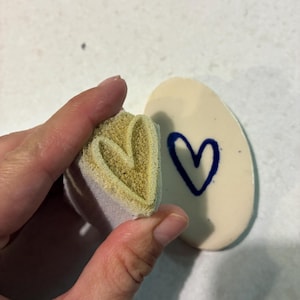 Op de afbeelding: Een beige schuimrubberen stempel met een hartvormig reliëfpatroon en een glad wit ovaal keramisch stuk met een donkerblauwe handgeschilderde hartomtrek. De stempel is zo geplaatst dat het hartontwerp overeenkomt met het blauwe hart op het keramiek.