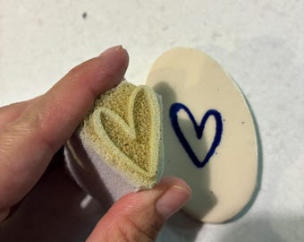 May include: A tan foam stamp with a heart-shaped relief pattern and a smooth white ceramic oval piece featuring a dark blue hand-painted heart outline. The stamp is held in position to show the heart design that matches the blue heart on the ceramic.