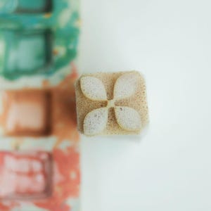 May include: A small, square, beige foam stamp featuring a four-petaled flower design carved into its surface. The stamp is positioned next to a blurred, multi-coloured paint palette containing squares of teal, peach, and pink watercolour pigments.