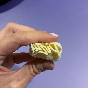 Può includere: Un piccolo timbro rettangolare in schiuma color bianco sporco con la parola "PIZZA" incisa in un carattere corsivo stilizzato e in rilievo. Il timbro ha una consistenza porosa e viene tenuto tra le dita, mostrando le lettere rovesciate.