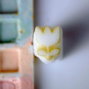 Puede incluir: Un pequeño bloque rectangular blanco con un diseño de tulipán amarillo estampado en su superficie frontal. El gráfico del tulipán está centrado y muestra una cabeza de flor simple con dos hojas en la base, en un pigmento amarillo dorado texturizado.