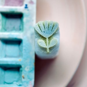 May include: A small, rectangular stamp featuring a carved flower design with a blue-tinted top and a green-tinted stem and leaves, positioned next to a rectangular paint palette containing multiple square wells of various blue and purple pigments.