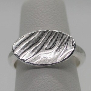 Puede incluir: Un anillo de plata con una parte superior ovalada que presenta un diseño abstracto texturizado.