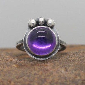 Puede incluir: Un anillo de plata con una amatista púrpura engastada en un engaste de bisel. El anillo tiene tres pequeñas bolas de plata en la parte superior de la banda.