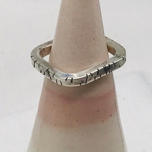 Puede incluir: Un anillo de plata con forma cuadrada y superficie texturizada. El anillo se muestra en un soporte de anillo de cerámica.