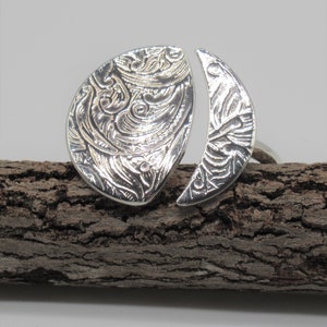 Puede incluir: Un anillo de plata con un diseño único. El anillo está hecho de dos piezas, una con forma de media luna y la otra con forma de lágrima. Ambas piezas están decoradas con intrincados patrones.