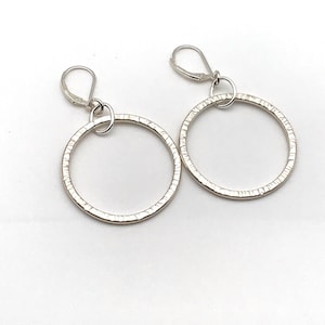 Puede incluir: Un par de pendientes de aro de plata con un acabado texturizado. Los pendientes tienen un diseño simple y moderno.