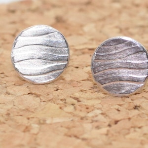Puede incluir: Un par de pendientes de plata con un patrón ondulado. Los pendientes son redondos y tienen un acabado liso y pulido.
