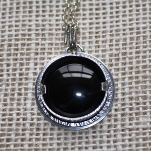 Puede incluir: Un collar de plata con un colgante de piedra preciosa negra engastado en un bisel redondo. El colgante tiene un borde texturizado.