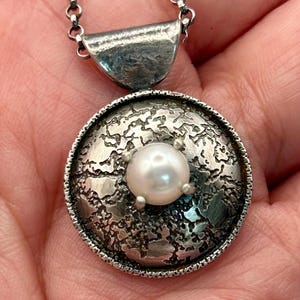 Puede incluir: Un collar con colgante de plata con un colgante circular texturizado y una perla blanca central. El colgante está unido a una delicada cadena. El diseño tiene un aspecto artesanal, con detalles intrincados alrededor de la perla.