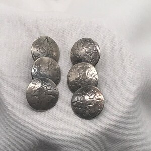 Puede incluir: Cinco pendientes redondos plateados con una superficie texturizada. Los pendientes están apilados uno encima del otro.