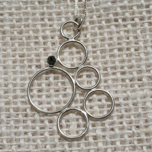 Puede incluir: Un collar de plata con un colgante que presenta un grupo de círculos interconectados, cada uno con un borde liso y redondeado. Una pequeña piedra preciosa negra está engastada en el centro del colgante.