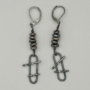 Puede incluir: Un par de pendientes plateados. Cada pendiente presenta una serie de pequeñas cuentas de color gris oscuro suspendidas de un cierre de palanca. Debajo de las cuentas hay un diseño envuelto en alambre con una forma geométrica.