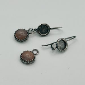 Puede incluir: Un par de pendientes de plata y un colgante a juego. Los pendientes presentan una pequeña piedra redonda de color marrón claro con un bisel plateado festoneado. El colgante tiene una piedra y un bisel similares.