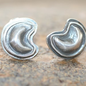 Puede incluir: Un par de pendientes de plata con un diseño de luna creciente. Los pendientes están hechos de metal texturizado y tienen una forma ligeramente redondeada.