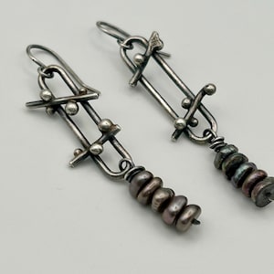 Puede incluir: Pendientes de plata hechos a mano con un diseño abstracto. Cada pendiente presenta barras y esferas de plata, así como una hilera de cuentas oscuras e iridiscentes. Cierre de gancho.