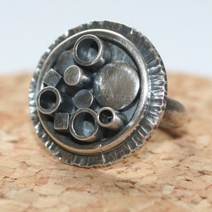 Puede incluir: Un anillo de plata con un diseño circular que presenta un borde exterior texturizado y un área central llena de varias formas geométricas, incluidos círculos, cuadrados y rectángulos.
