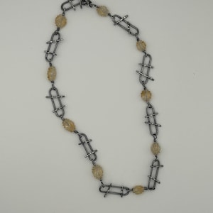 Puede incluir: Un collar con una serie de eslabones de metal plateado intercalados con cuentas texturizadas de color amarillo claro. El collar tiene un cierre de palanca y se muestra sobre un fondo blanco. La joya es un collar.