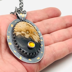 Puede incluir: Un collar con un colgante de plata y oro con una piedra de ágata negra y marrón con un acento de piedra preciosa amarilla. El colgante está rodeado por un borde decorativo de plata con acentos dorados.
