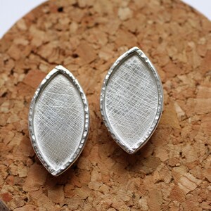 Puede incluir: Dos componentes de joyería plateados de forma ovalada. Cada componente presenta una superficie de metal cepillado texturizado dentro de un marco con borde martillado. Los componentes se muestran sobre un fondo de corcho.