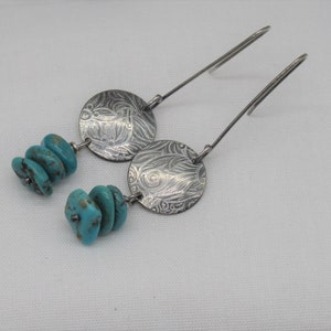 Puede incluir: Pendientes colgantes plateados con detalles de piedras de color turquesa. Cada pendiente tiene un diseño circular grabado y una pila de tres piedras turquesas. Los pendientes tienen ganchos largos y finos.