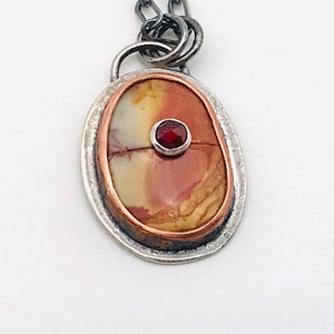 Puede incluir: Un colgante de plata y cobre con una piedra preciosa roja engastada en una piedra con patrones marrones, blancos y naranjas.