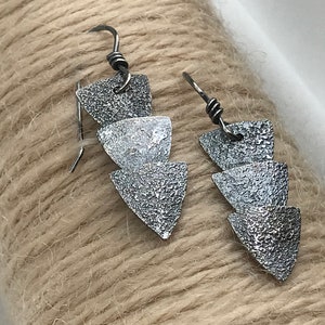 Puede incluir: Un par de pendientes de plata con un diseño triangular texturizado. Cada pendiente presenta tres triángulos apilados, creando un aspecto geométrico y moderno.