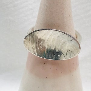 Puede incluir: Un anillo de plata con una parte superior de forma ovalada. La parte superior del anillo tiene un diseño texturizado y martillado.