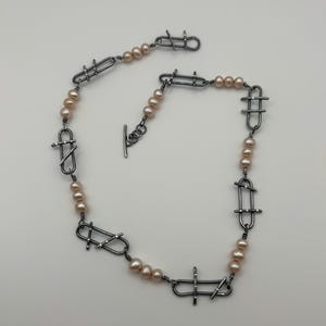 Puede incluir: Un collar con perlas color rubor y metal plateado. El diseño presenta eslabones metálicos geométricos entre las perlas. El cierre es de estilo palanca. La joya se exhibe sobre un fondo liso. Un collar único.