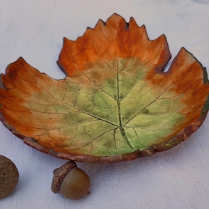 Könnte beinhalten: Eine flache, blattförmige Schale mit einem Farbverlauf in Herbsttönen, der von einem leuchtend grünen Zentrum zu tief orangen und braunen Rändern übergeht. Die Oberfläche zeigt detaillierte, geprägte Aderstrukturen und zwei kleine braune Eicheln liegen daneben.
