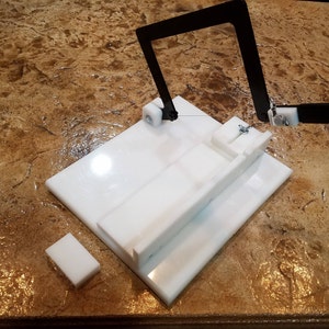 May include: A white plastic cheese cutting tool with a black metal frame and a wire for cutting. The tool is on a brown countertop.